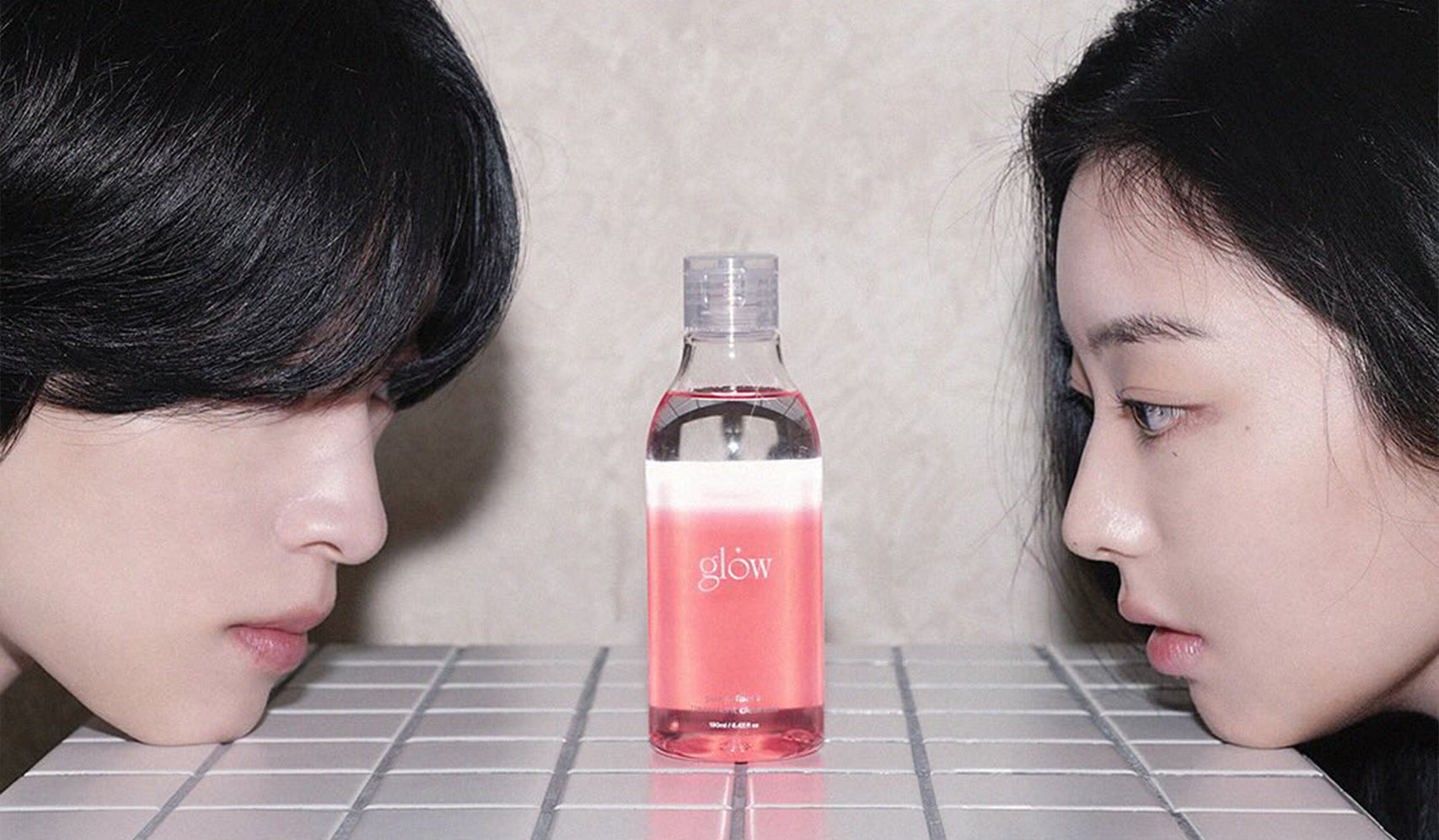 Two people staring at a bottle of glow skincare product on a tiled surface.