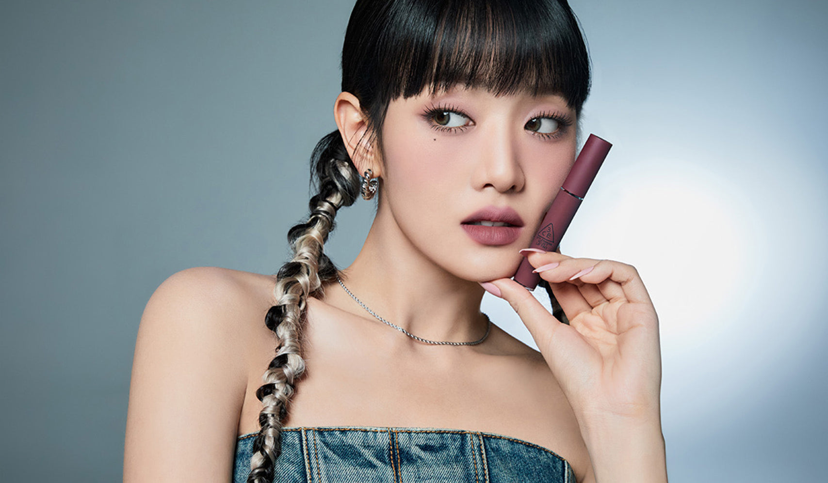 Woman with braided hair holding a lipstick against a gray background, showcasing beauty and skincare elegance.