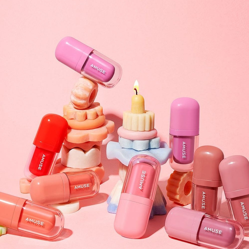 Colorful arrangement of AMUSE BEBE TINT lip tints with candle display against pink background.