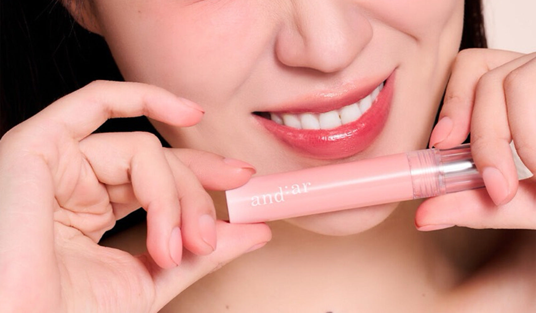 Close-up of person holding a tube of pink lip gloss while smiling, showcasing shiny lips and dental care.