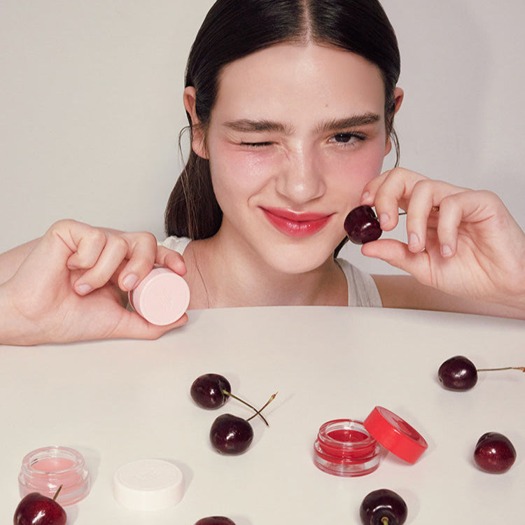 Woman holding ANILLO Mellow Glow Balm with cherries and balm containers on table.