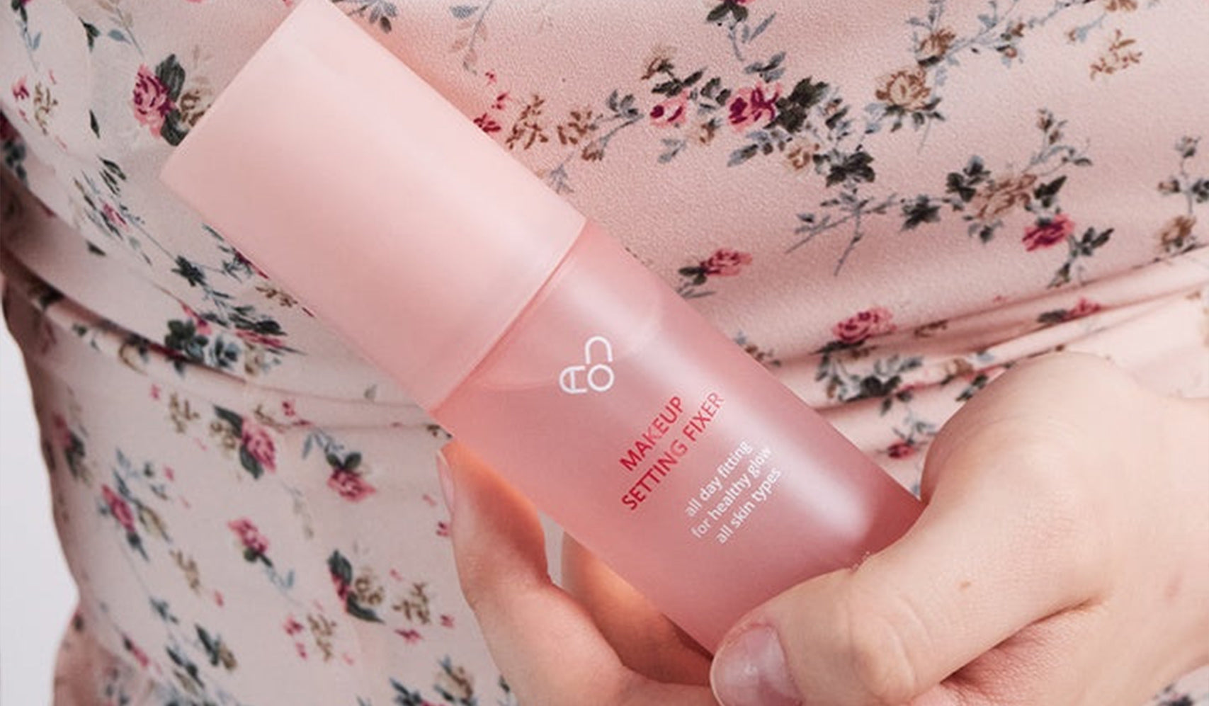Person holding a pink makeup setting spray bottle, wearing a floral-patterned dress.