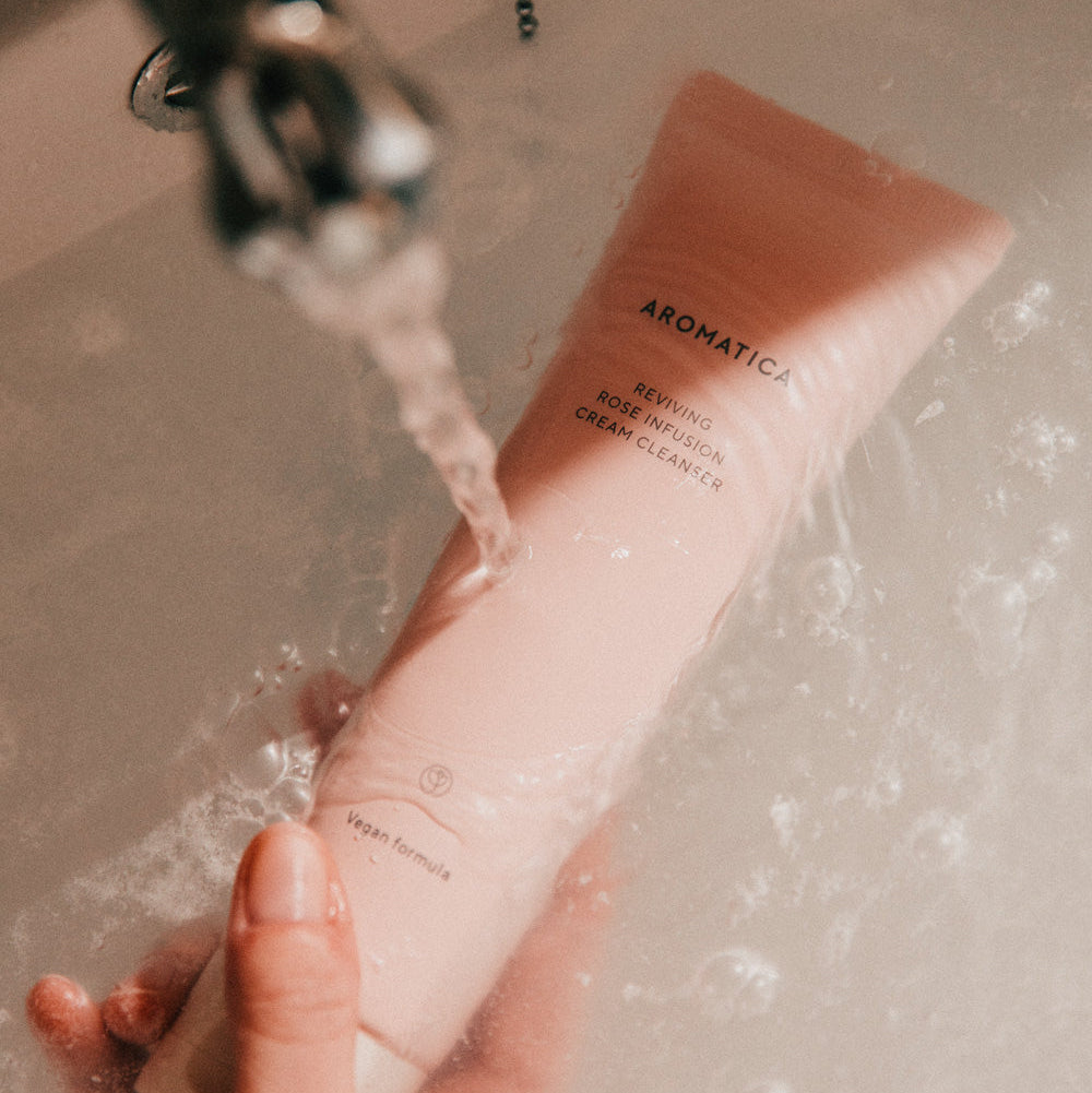 Hand holding AROMATICA Reviving Rose Infusion Cream Cleanser 145ML under running water
