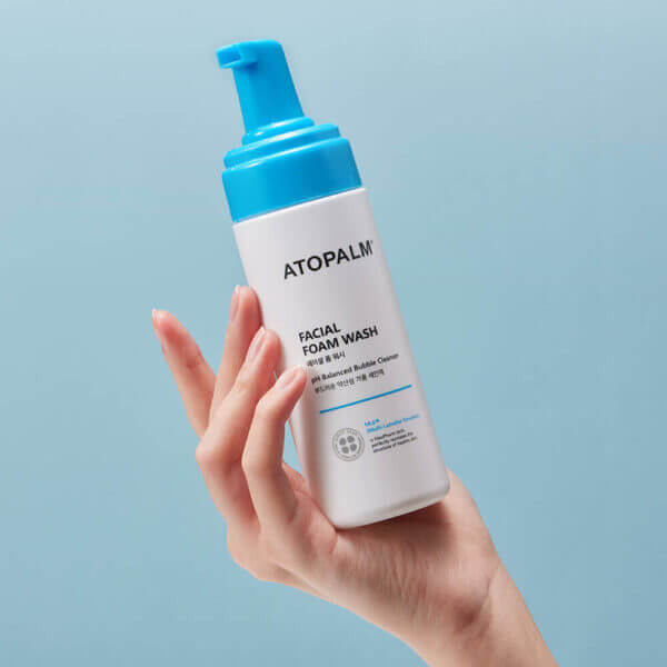 Hand holding ATOPALM Facial Foam Wash 150ml bottle against blue background