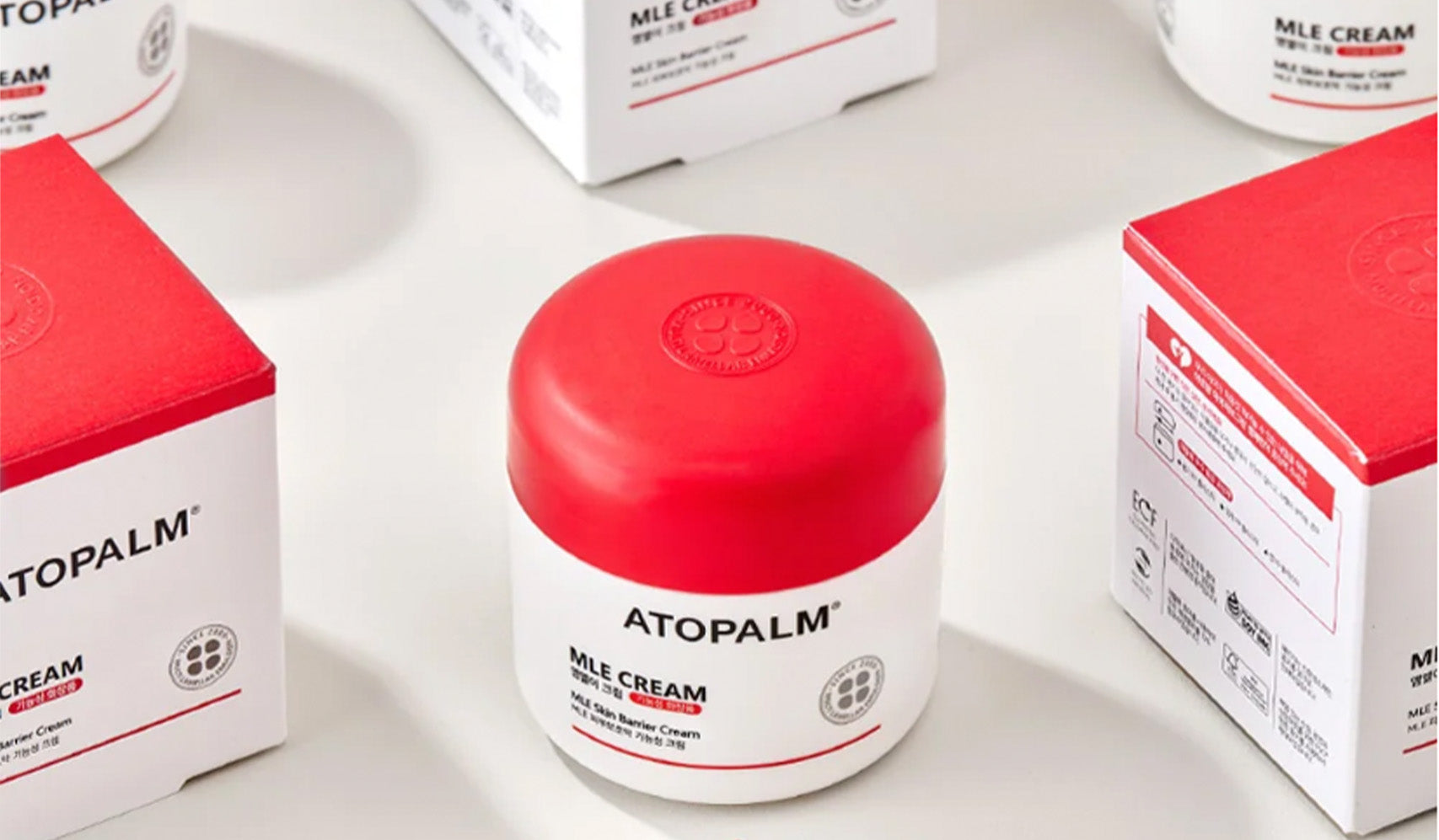 Atopalm MLE Cream jar with red lid surrounded by packaging on a light background