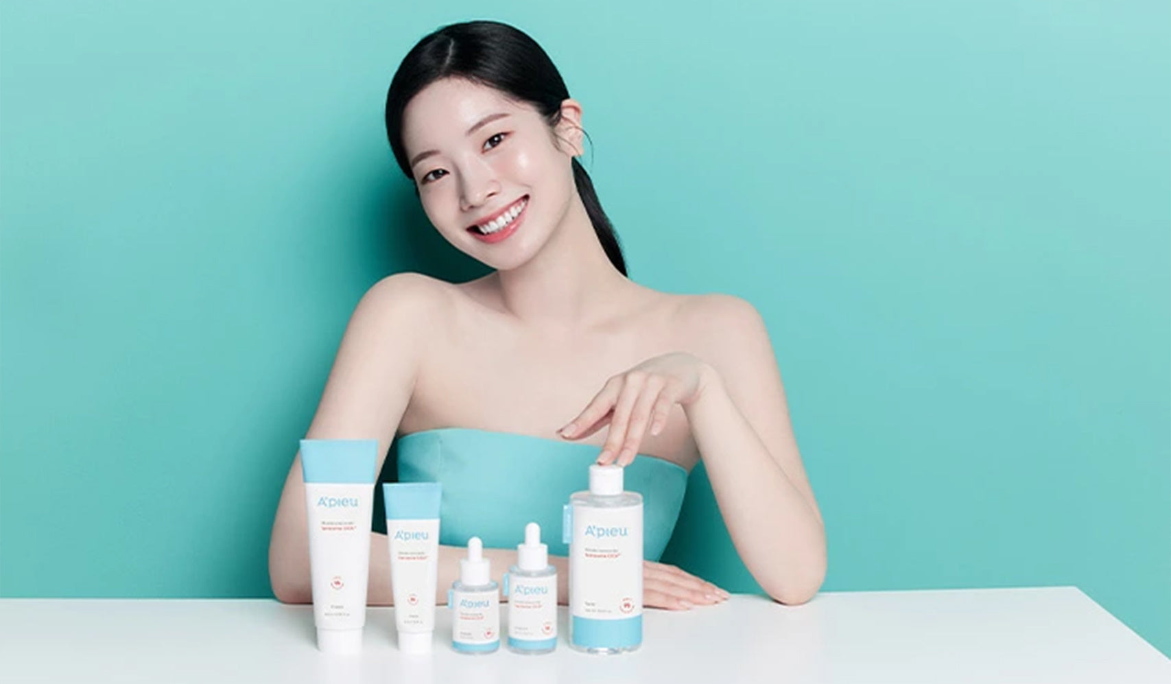 Smiling woman showcasing skincare products against a teal background, featuring a range of bottles and tubes for beauty care.