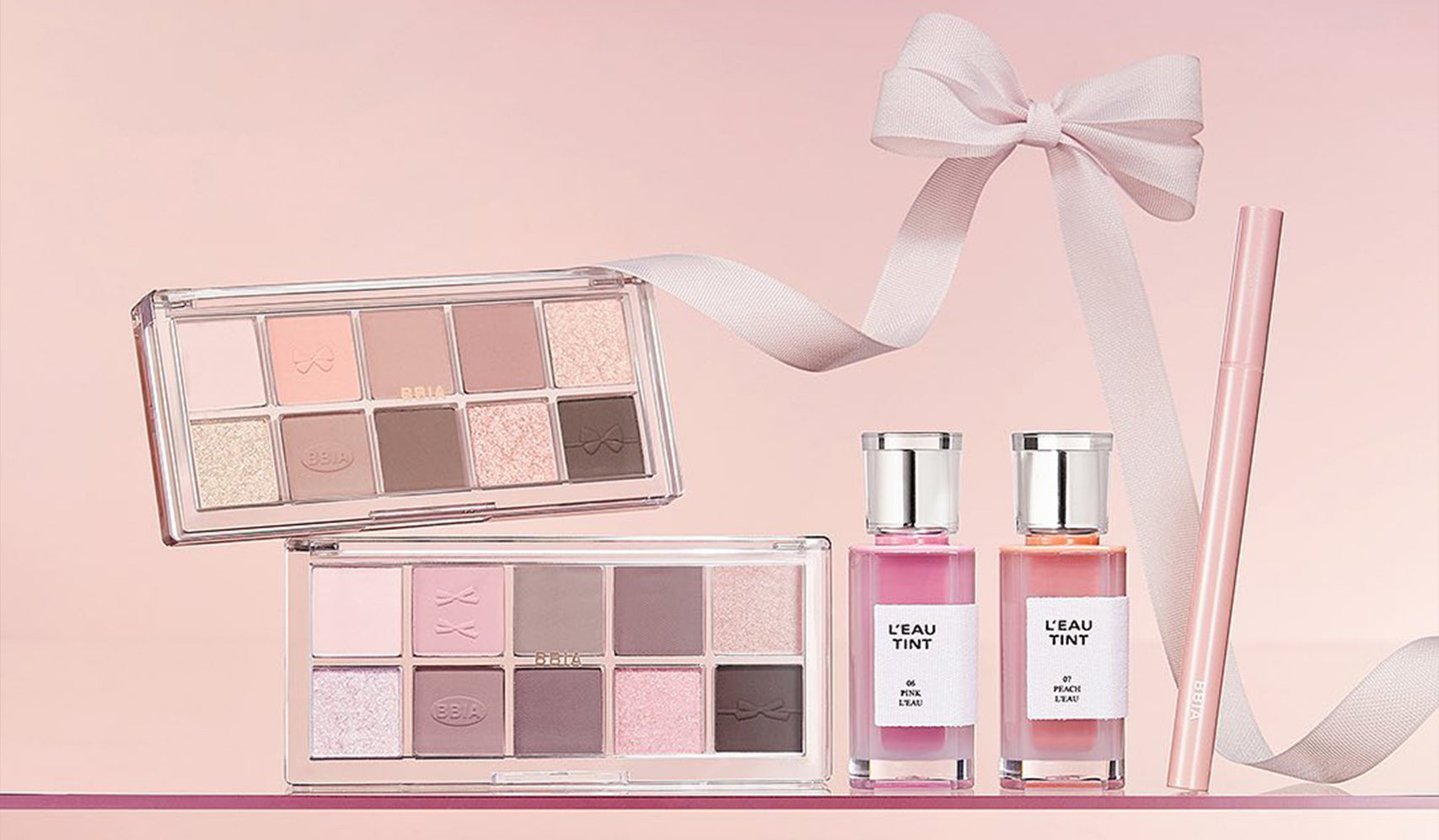Makeup set with two eyeshadow palettes and tinted water bottles on a pink background, featuring a ribbon decoration.