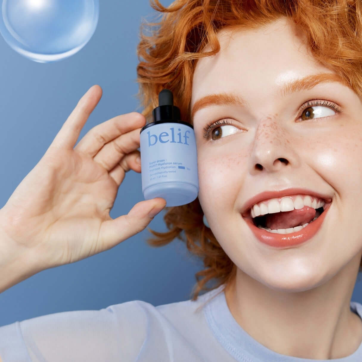 Woman holding BELIF Super Drops Multi 7-Hyaluron Serum 30ml against a blue background, showcasing skincare product.
