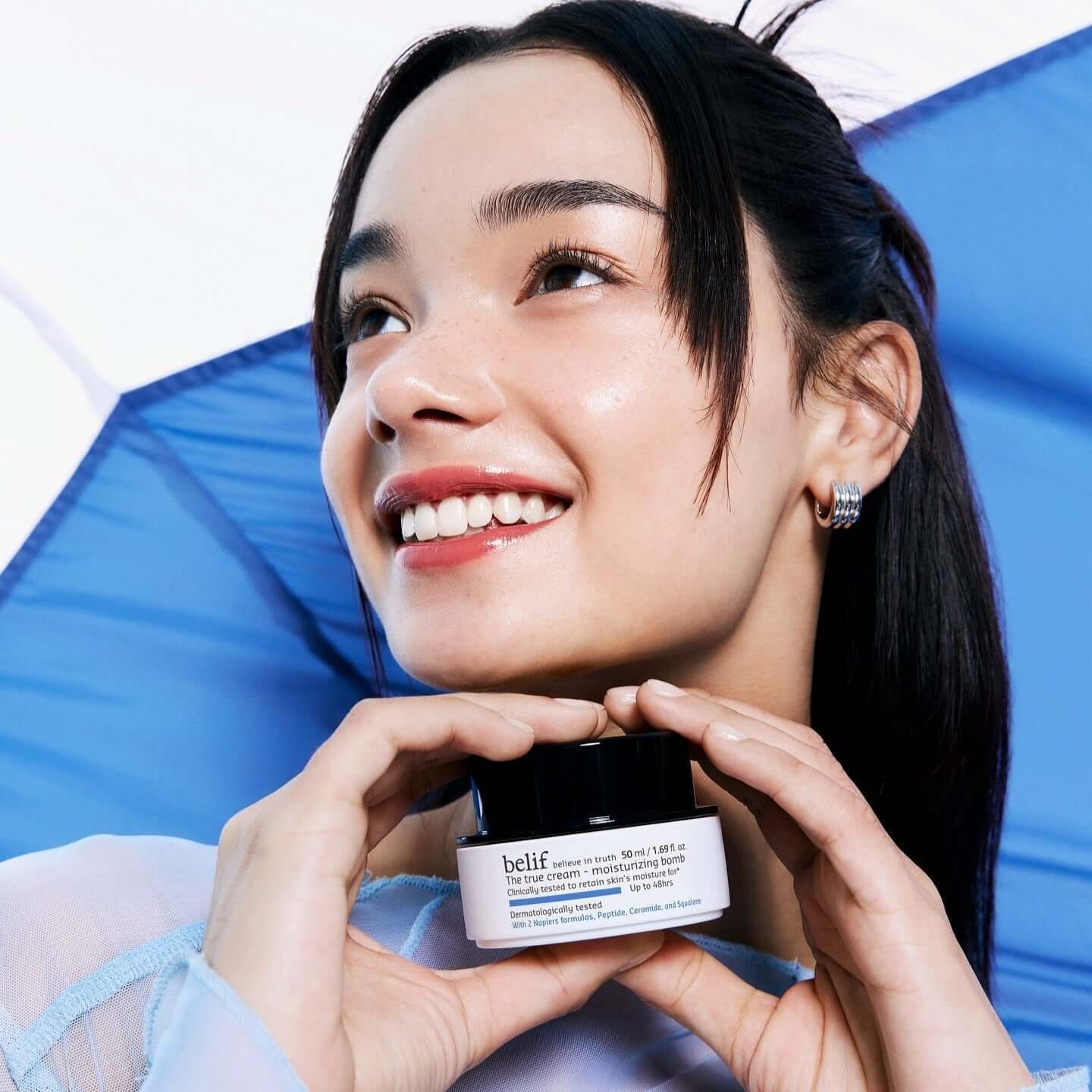 Smiling person holding BELIF The True Cream Moisturizing Bomb 50ml against a blue backdrop.