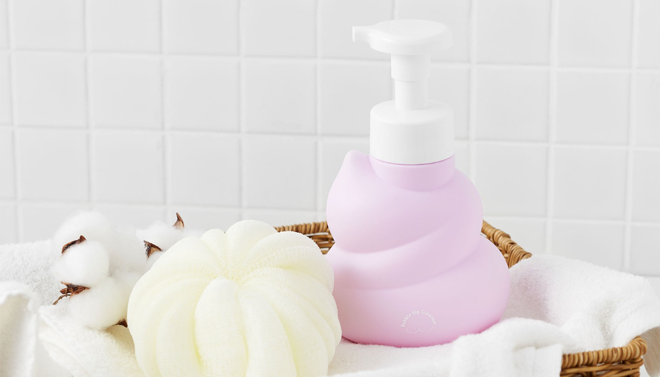 Pastel pink soap dispenser with puff and cotton balls in a basket, on a white tiled background.