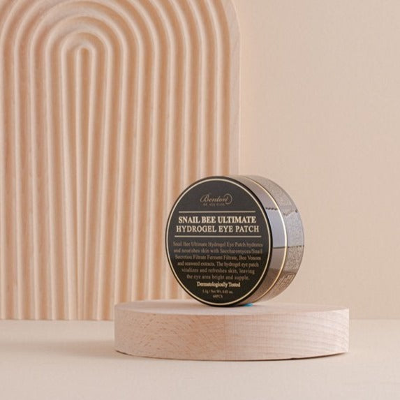 Benton Snail Bee Ultimate Hydrogel Eye Patch 70g container displayed on a wooden surface with a decorative backdrop.