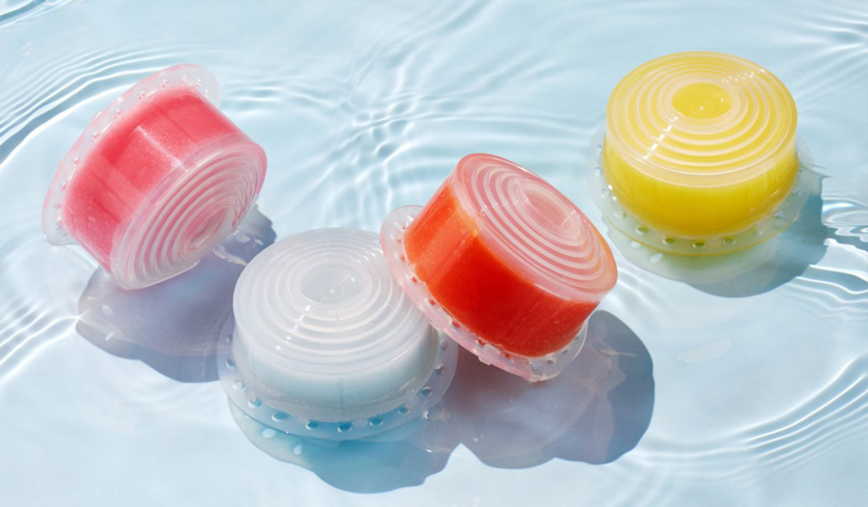 Colorful bath salts containers floating in clear water, featuring pink, white, red, and yellow boxes with circular lids.