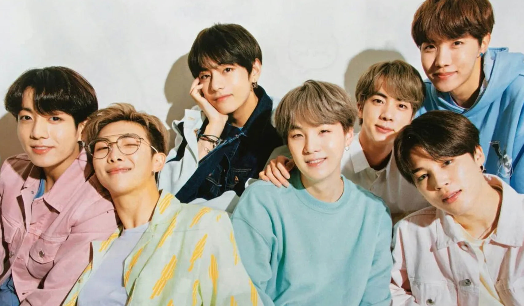 Group of seven men smiling together in colorful casual clothing against a neutral background.