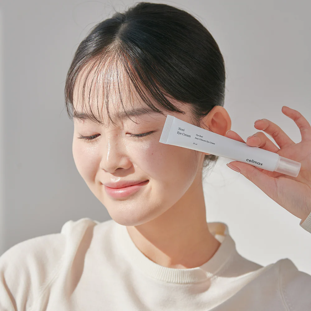 Woman holding CELIMAX The Real Noni Ultimate Eye Cream 20ml with a smile, showcasing skincare product.