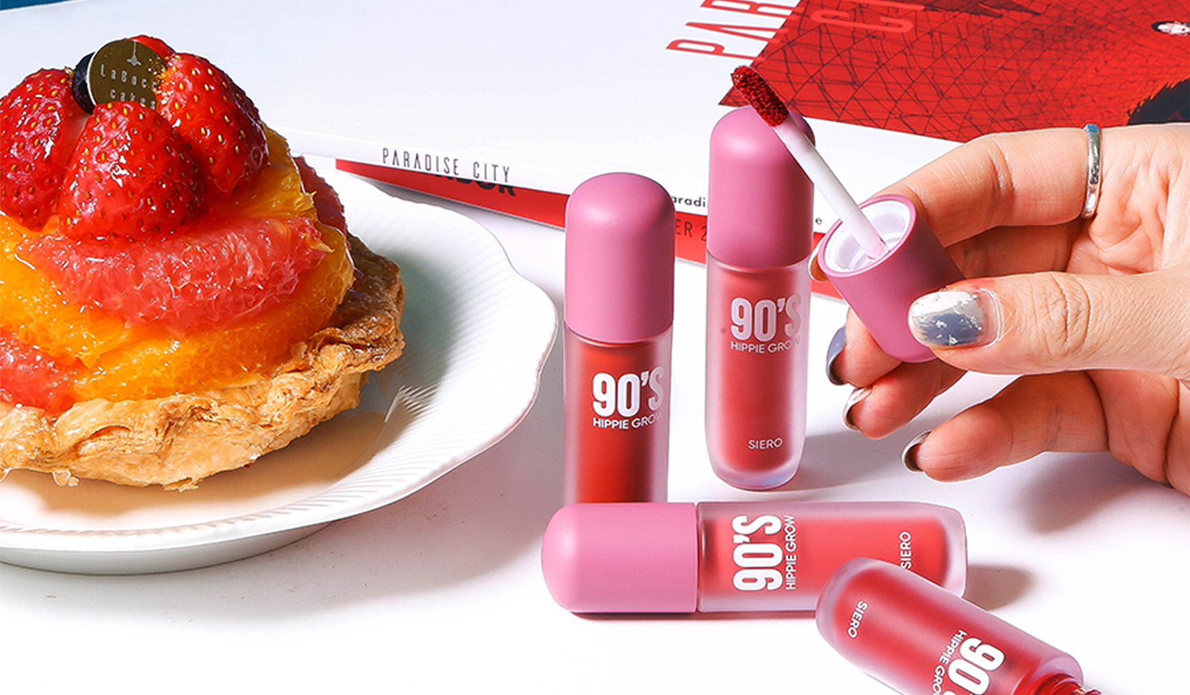 Lip tint set with pink packaging on a table beside a fruit tart and a red pencil, promoting '90s Hippie City' collection.