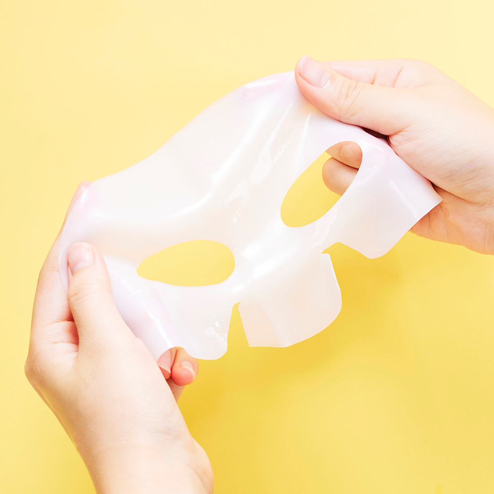 Hands holding COSRX Advanced Snail Mucin Glass Glow Hydrogel Mask (3 mask) against a yellow background.