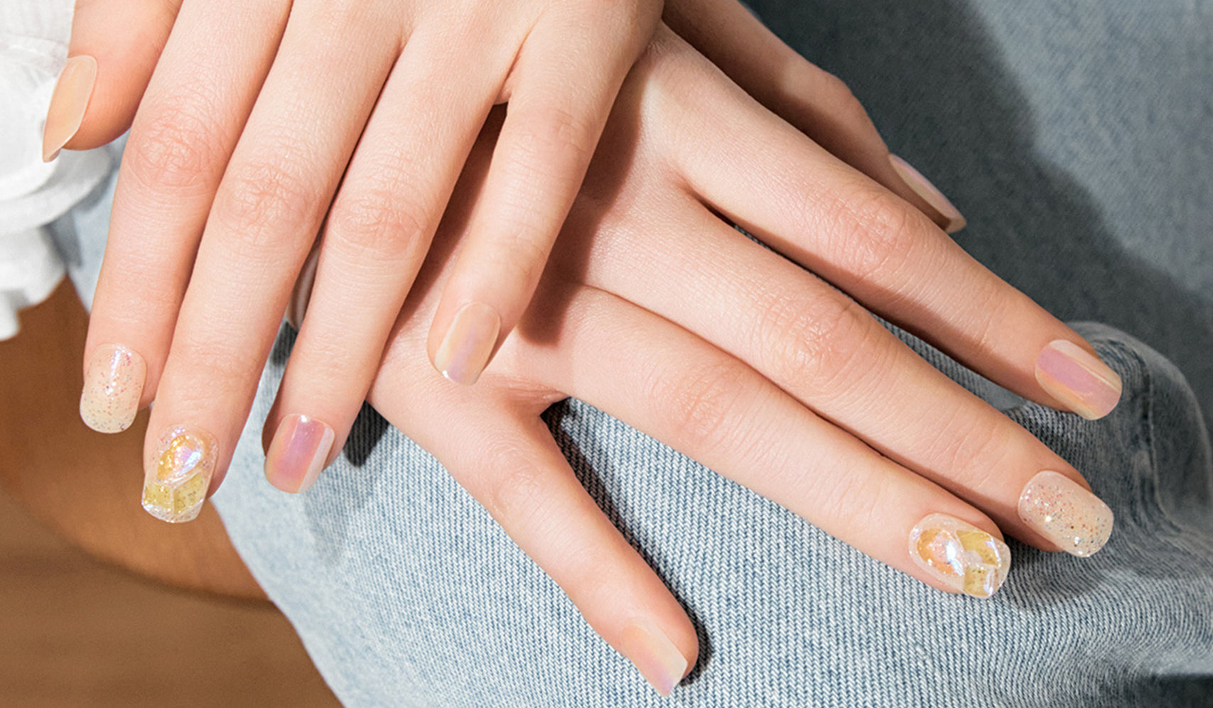 Person with artistic nail art featuring pastel colors and glitter on manicured nails, placed on denim fabric.