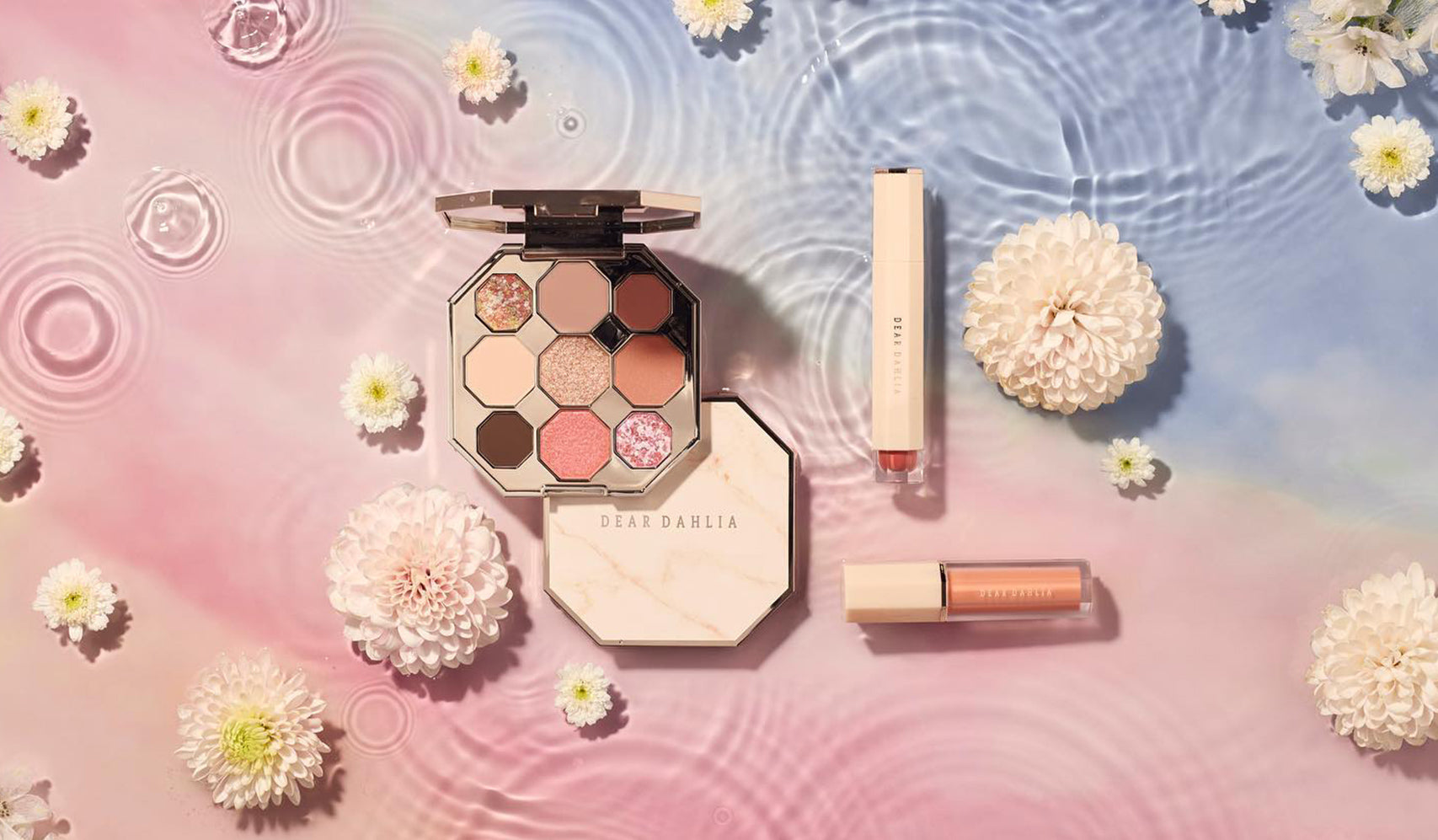 Delicate makeup set with palette, lipstick, and lip gloss on a pastel pink and blue water ripple background with flowers.