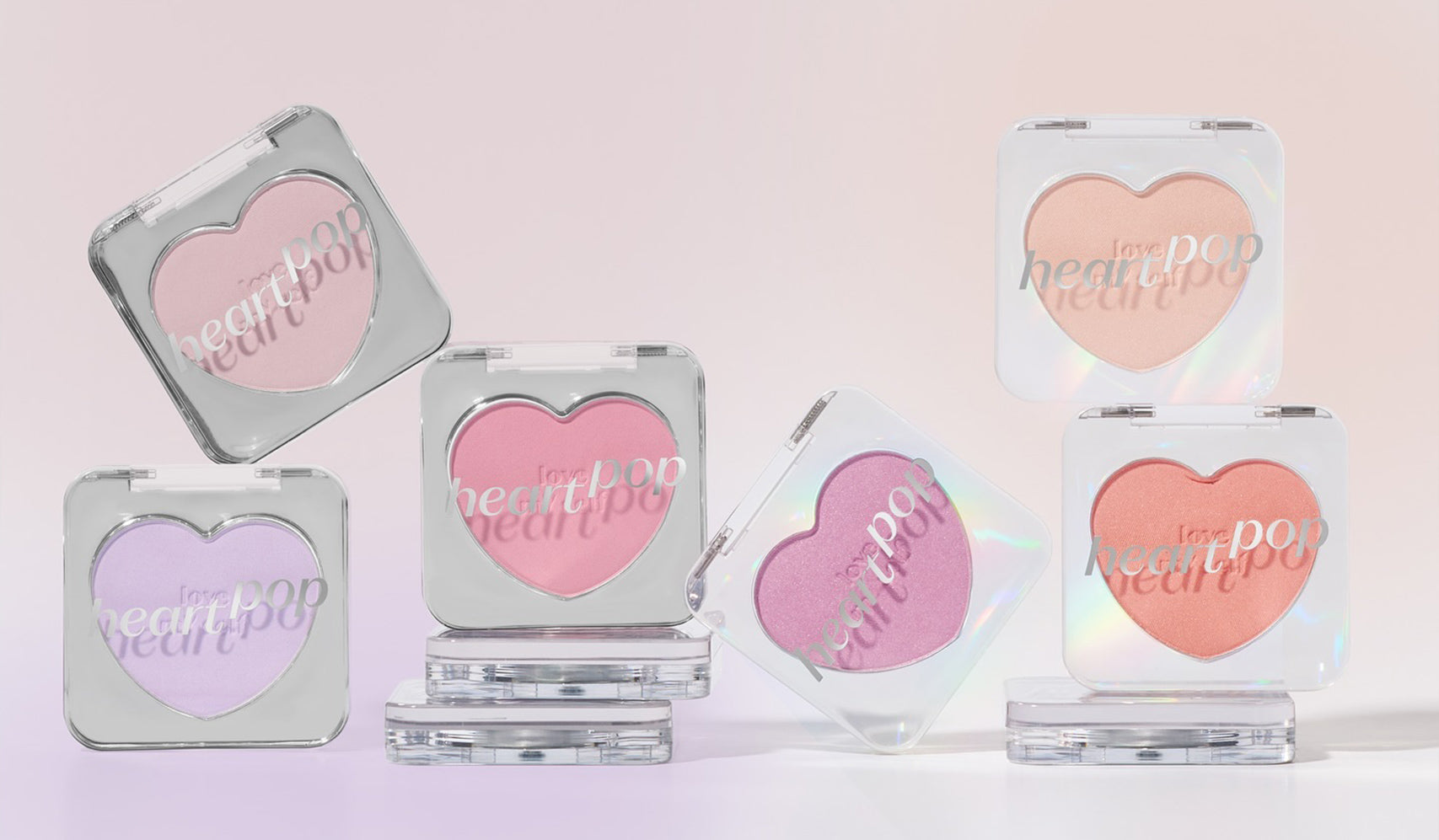 Heart-shaped pastel blush compacts in various shades on a soft pink background, showcasing cosmetic product packaging.