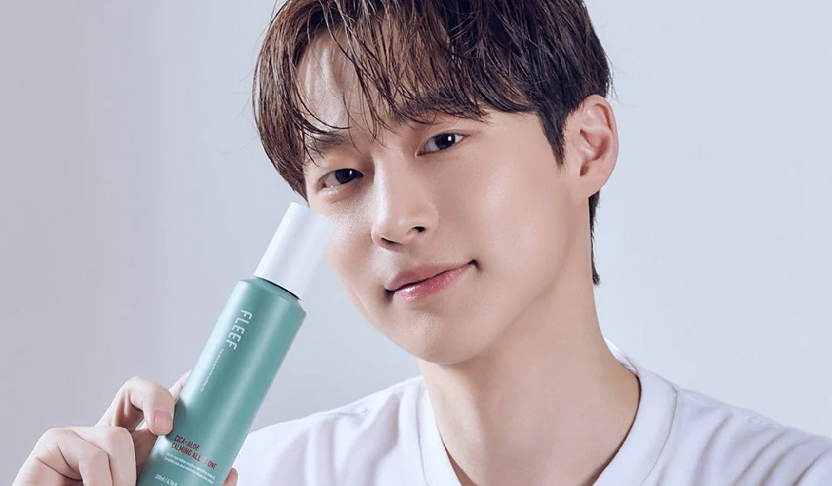 Person holding green skincare bottle, wearing a white shirt with a neutral background.