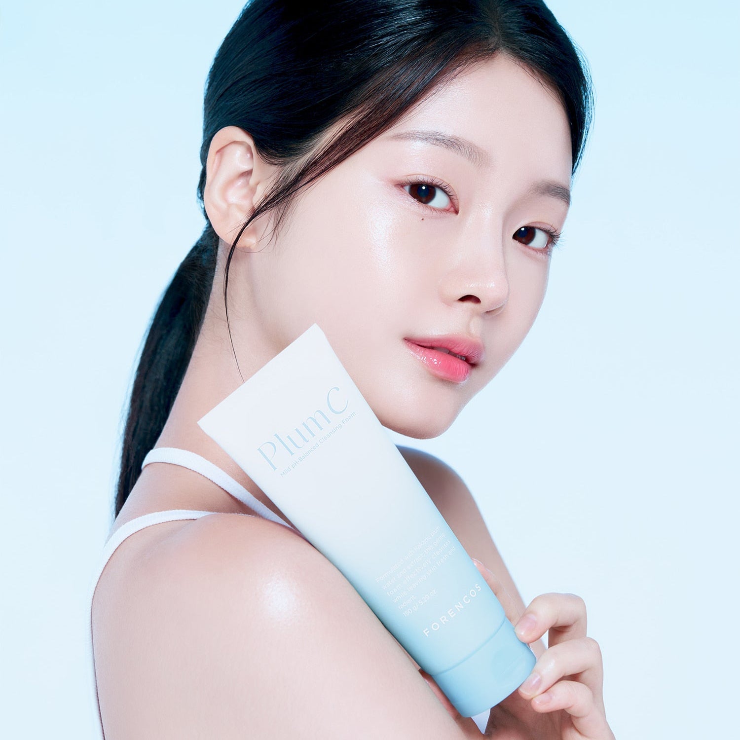 Woman holding FORENCOS Plum C Mild Mild Acidic Cleansing Foam 150g against blue background, showcasing skincare product.