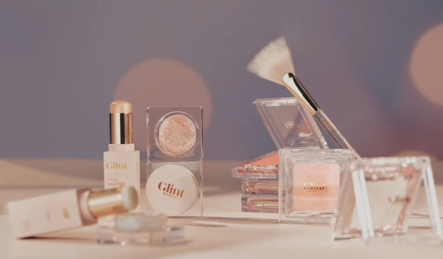 Assorted Glint beauty products including lipsticks, eyeshadow, and blush with soft lighting and a makeup brush on display.