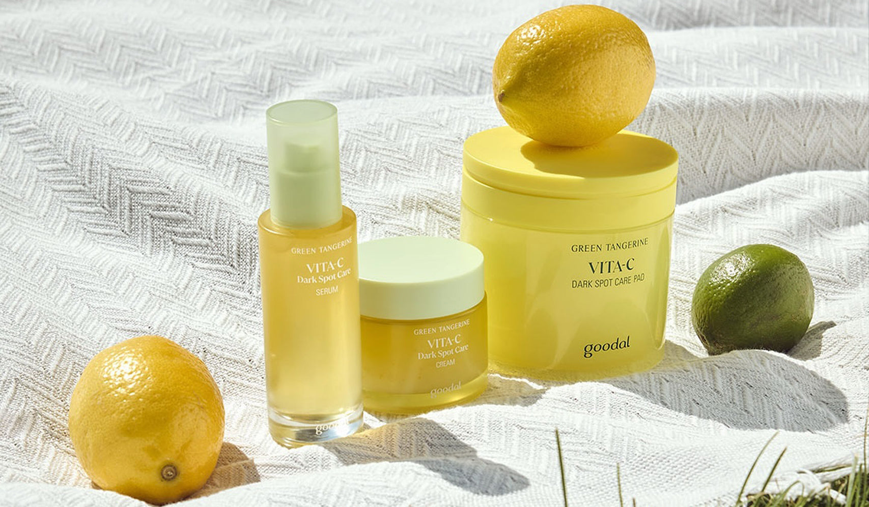 Green Tangerine Vita C skincare products with fresh lemons and limes on a white textured cloth.