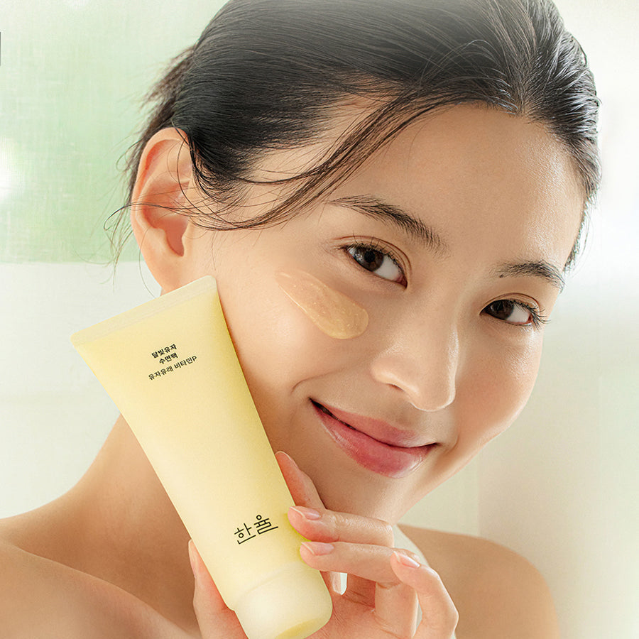 Woman applying HANYUL Moonlight Yuja Sleeping Pack 100ml on cheek, showcasing the tube and texture of the skincare product.