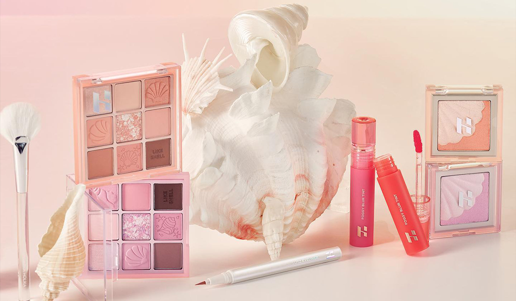 Makeup collection featuring eye shadow palettes, lip tint, and brushes with seashell decoration on a soft pink background.