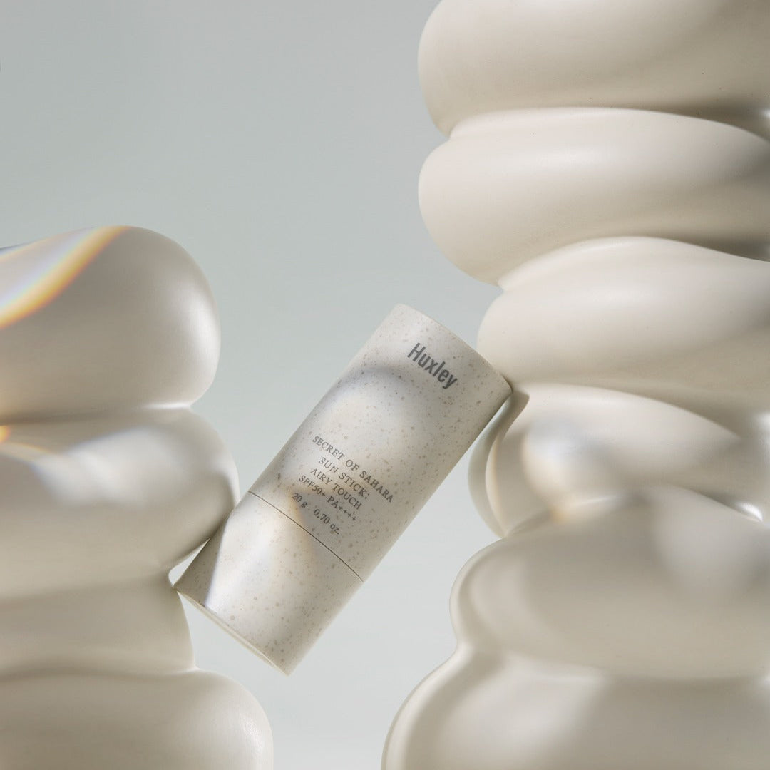 HUXLEY Sun Stick Airy Touch 20g in elegant packaging, surrounded by abstract shapes, offering UV protection and skincare benefits.
