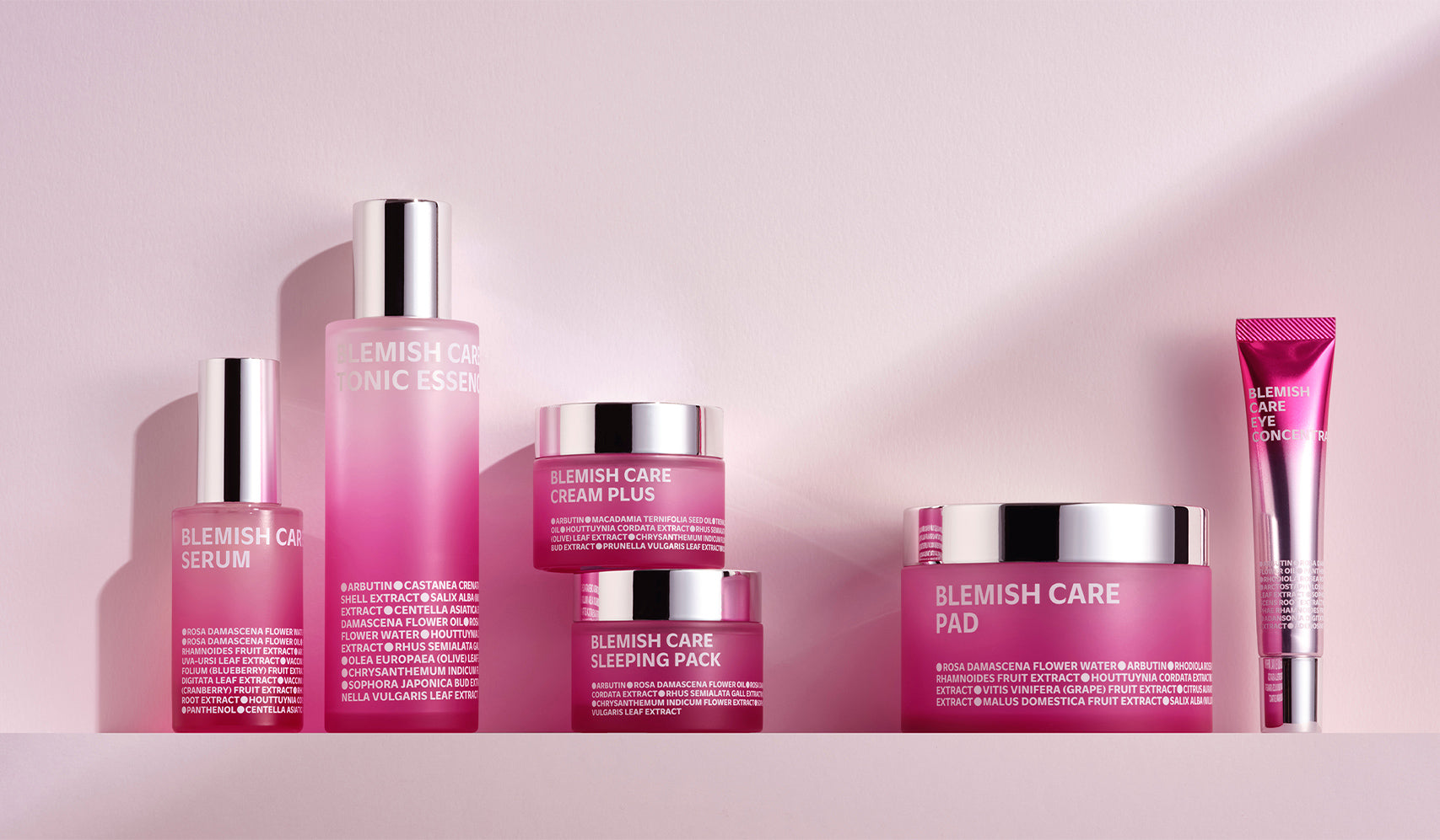 Collection of pink blemish care skincare products including serum, tonics, cream, and pads on a soft pink background.