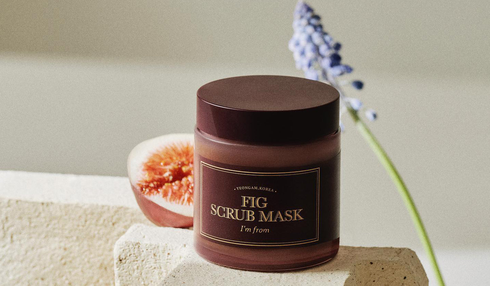Jar of "I'm From Fig Scrub Mask" with fig and flower in background, capturing a natural skincare setting.