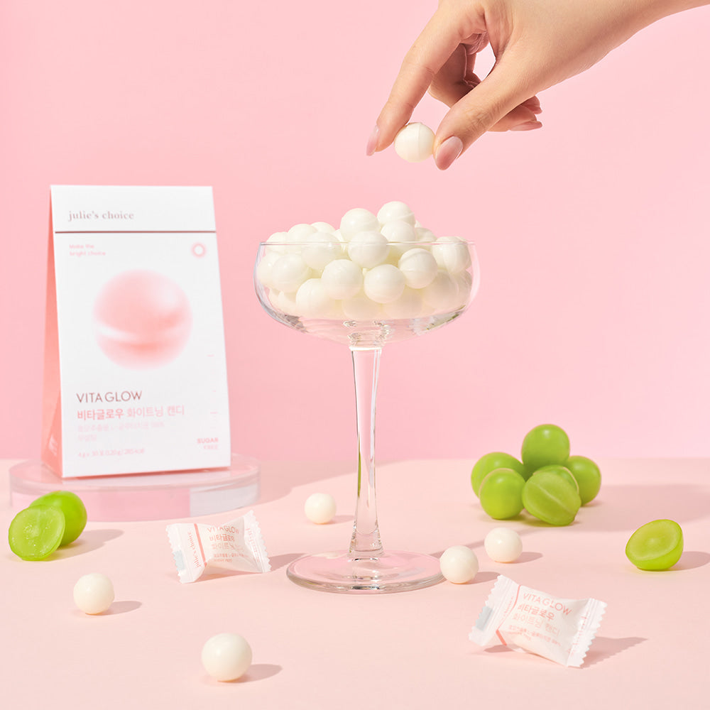 JULIE'S CHOICE Vita Glow Whitening Candy displayed in a glass with packaging and grapes on a pink background