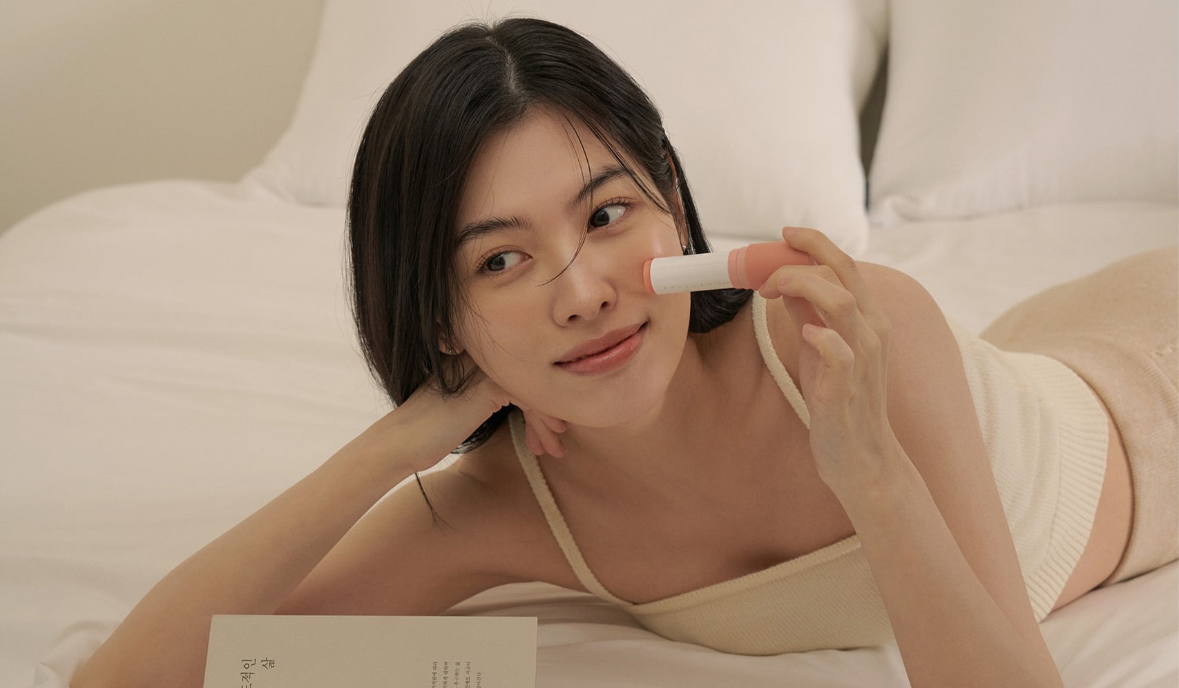 Woman relaxing on a bed, holding a skincare product, looking content