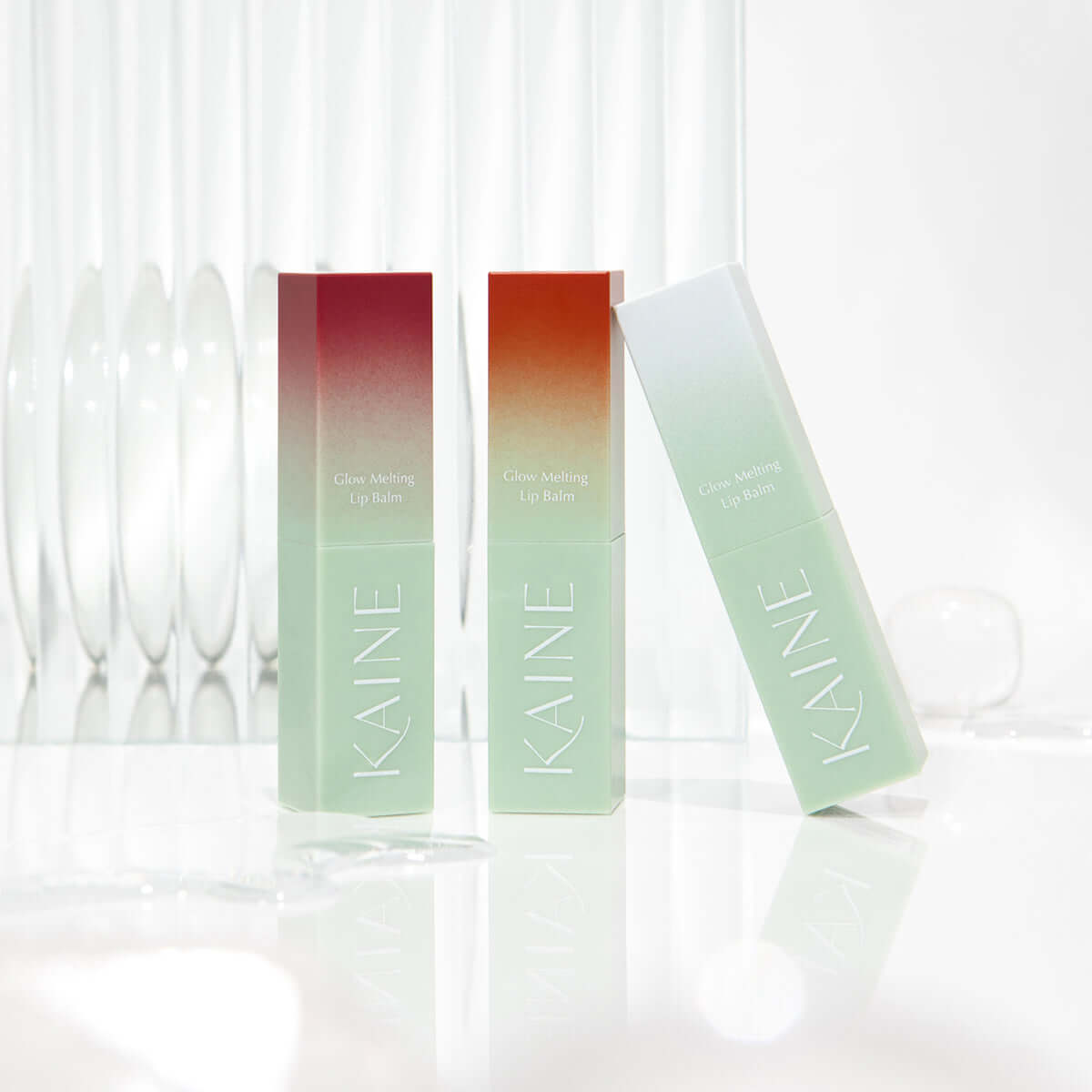 Three KAINE Glow Melting Lip Balm tubes standing on a reflective surface with a clear glass backdrop.