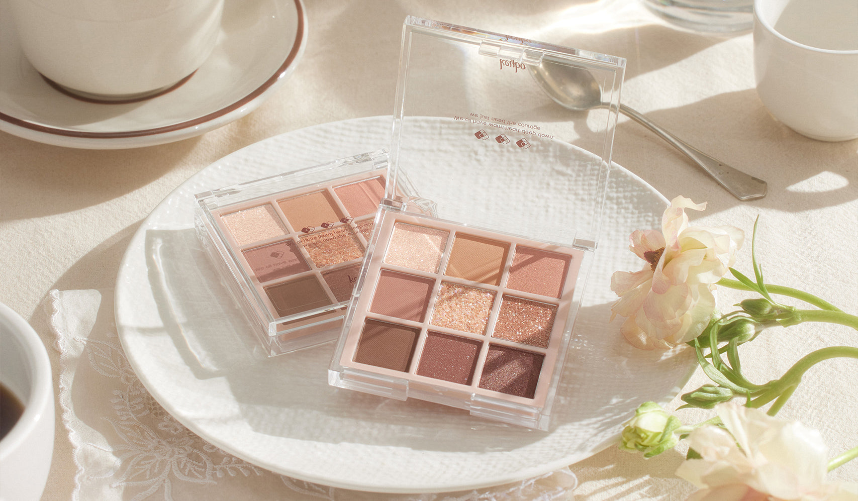 Eyeshadow palettes with neutral shades on a plate, surrounded by flowers and a cup, styled on a sunny table.