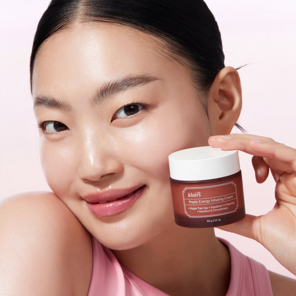 Smiling woman holding KLAIRS Maple Energy Infusing Cream 60g against light background.