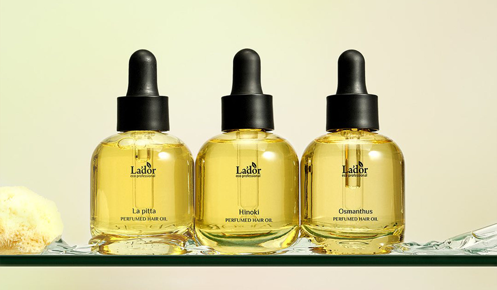 Three bottles of perfumed hair oil with dropper lids on a glass shelf, featuring La'dor, La Pitta, and Osmanthus labels.