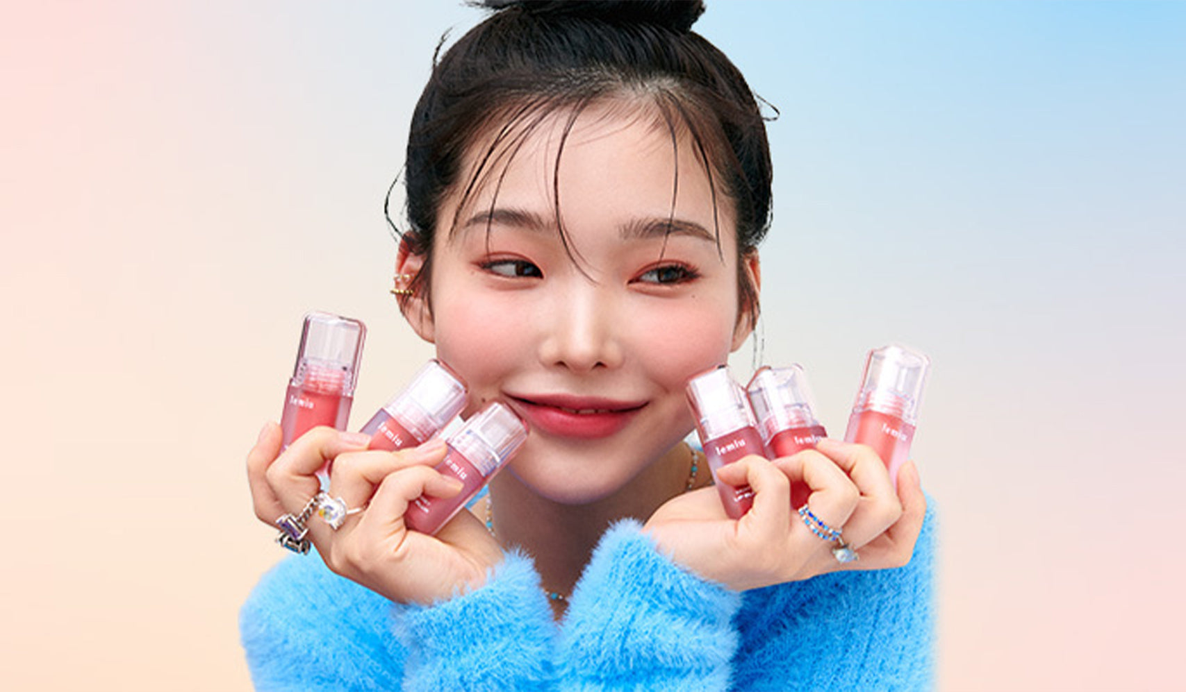 Person holding multiple lip gloss tubes, wearing a soft blue sweater in a vibrant, colorful background.