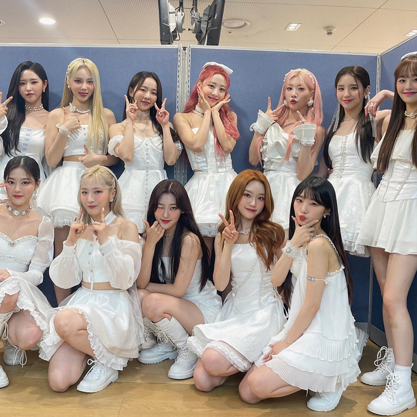 Group of people posing in white outfits, standing and sitting, making peace signs, indoors.