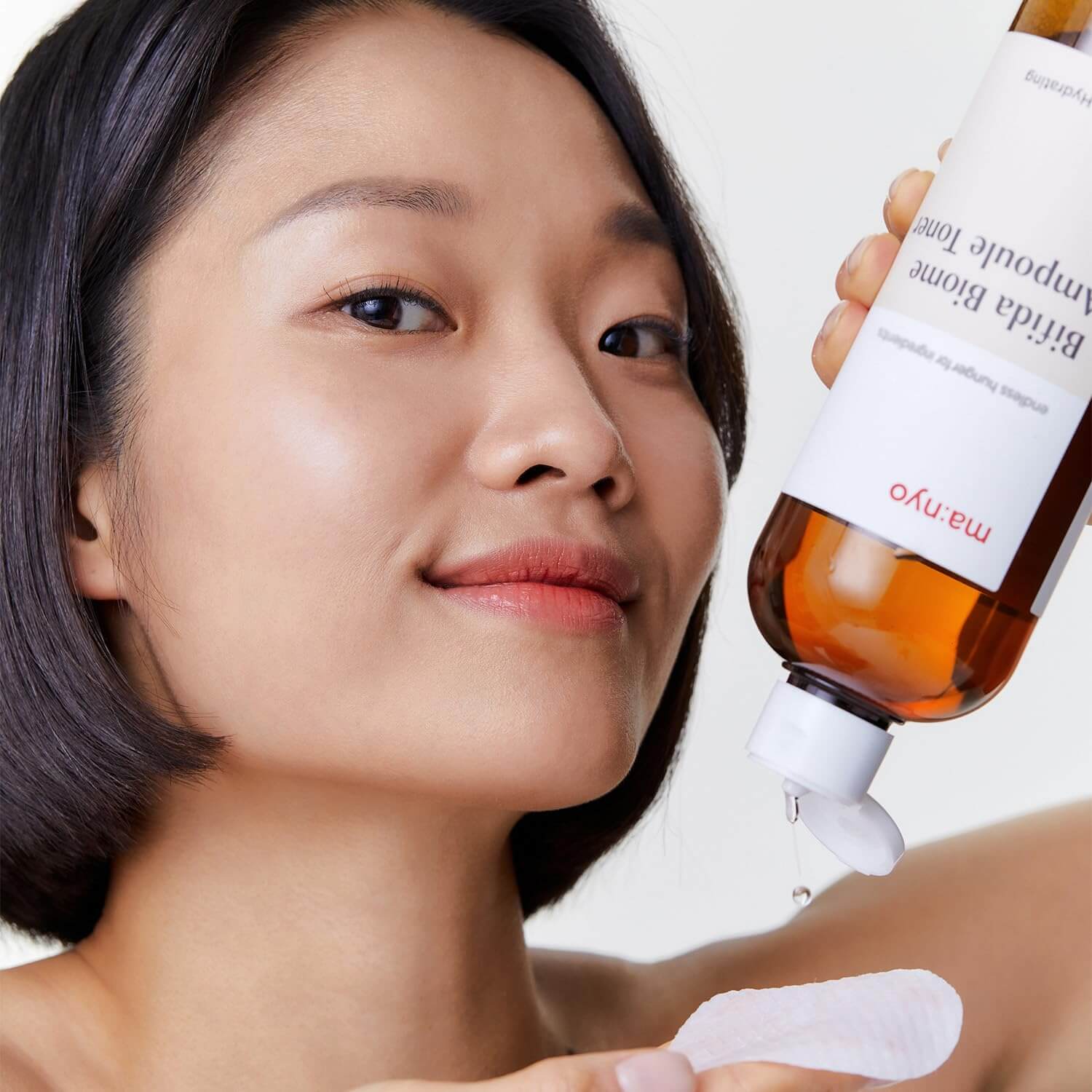 Woman applying MANYO Bifida Biome Ampoule Toner 400ml to cotton pad, showcasing skincare routine.