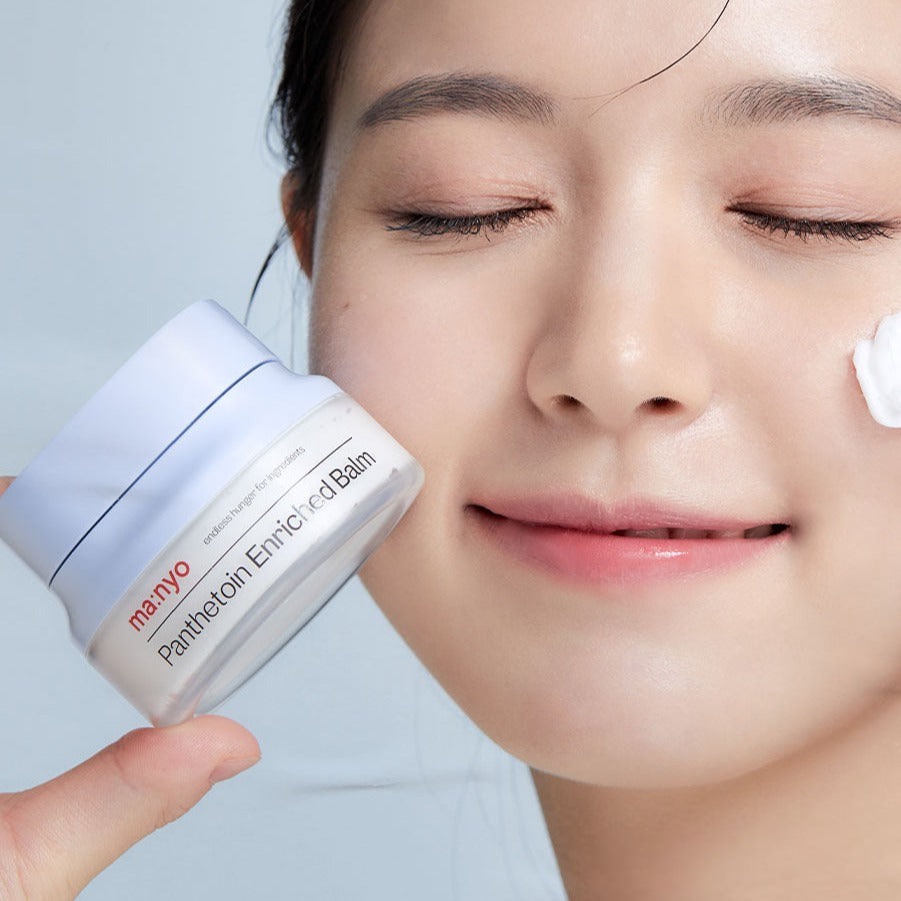 A woman applying MA:NYO Panthetoin Enriched Balm 80ml to her cheek, holding the product jar with a smile.