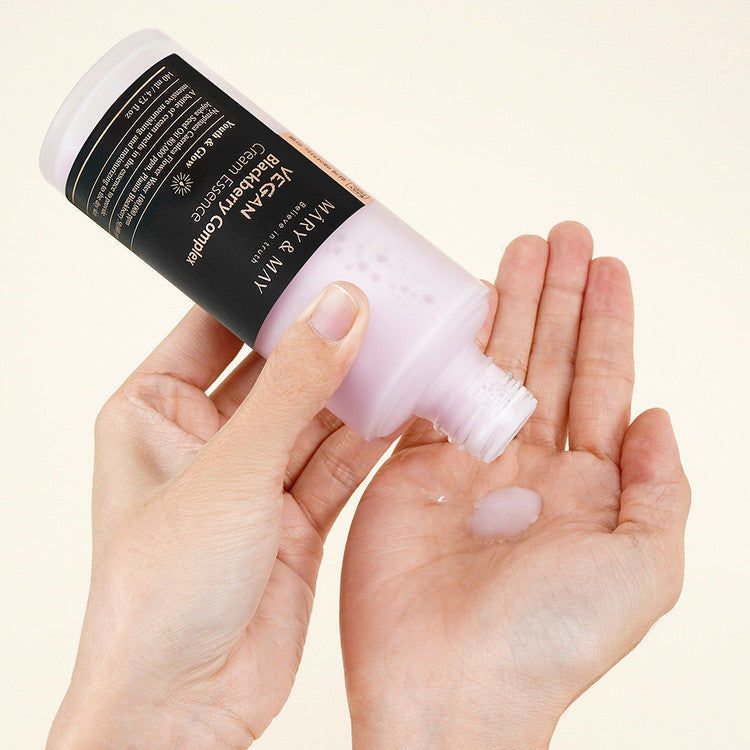 Hands holding MARY & MAY Vegan Blackberry Complex Cream Essence 40ml, pouring essence onto palm, showcasing texture and packaging.