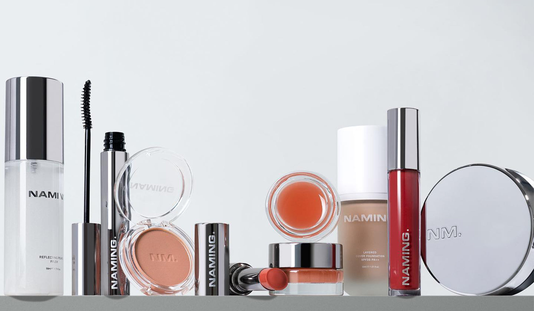 Selection of makeup products including mascara, blush, lip gloss, and foundation, displayed on a shelf with silver packaging.