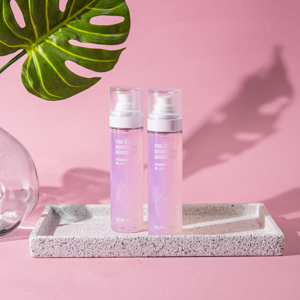 Two bottles of NEOGEN Pink Vita Aurora Mist 115ml on a tray with a palm leaf and pink background, showcasing product design.
