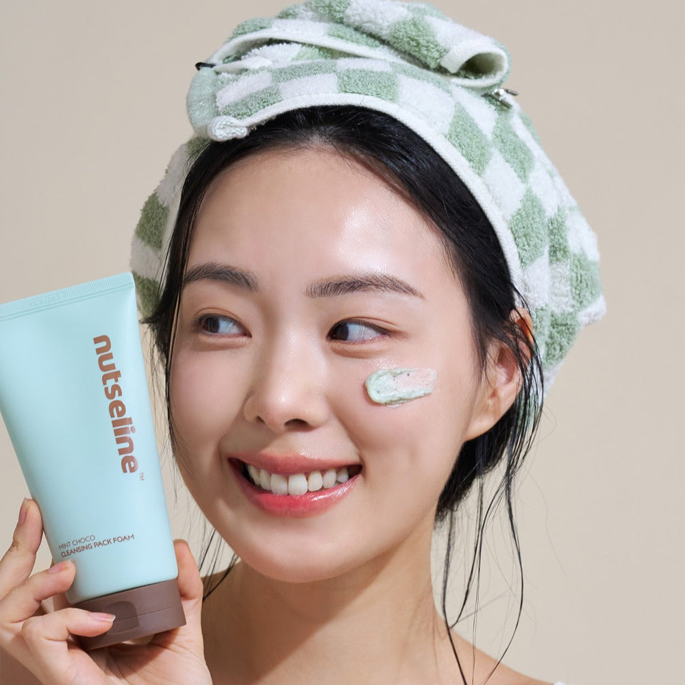 Smiling woman applying NUTSELINE Mint Chocolate Cleansing Pack Foam 130g with towel on head.