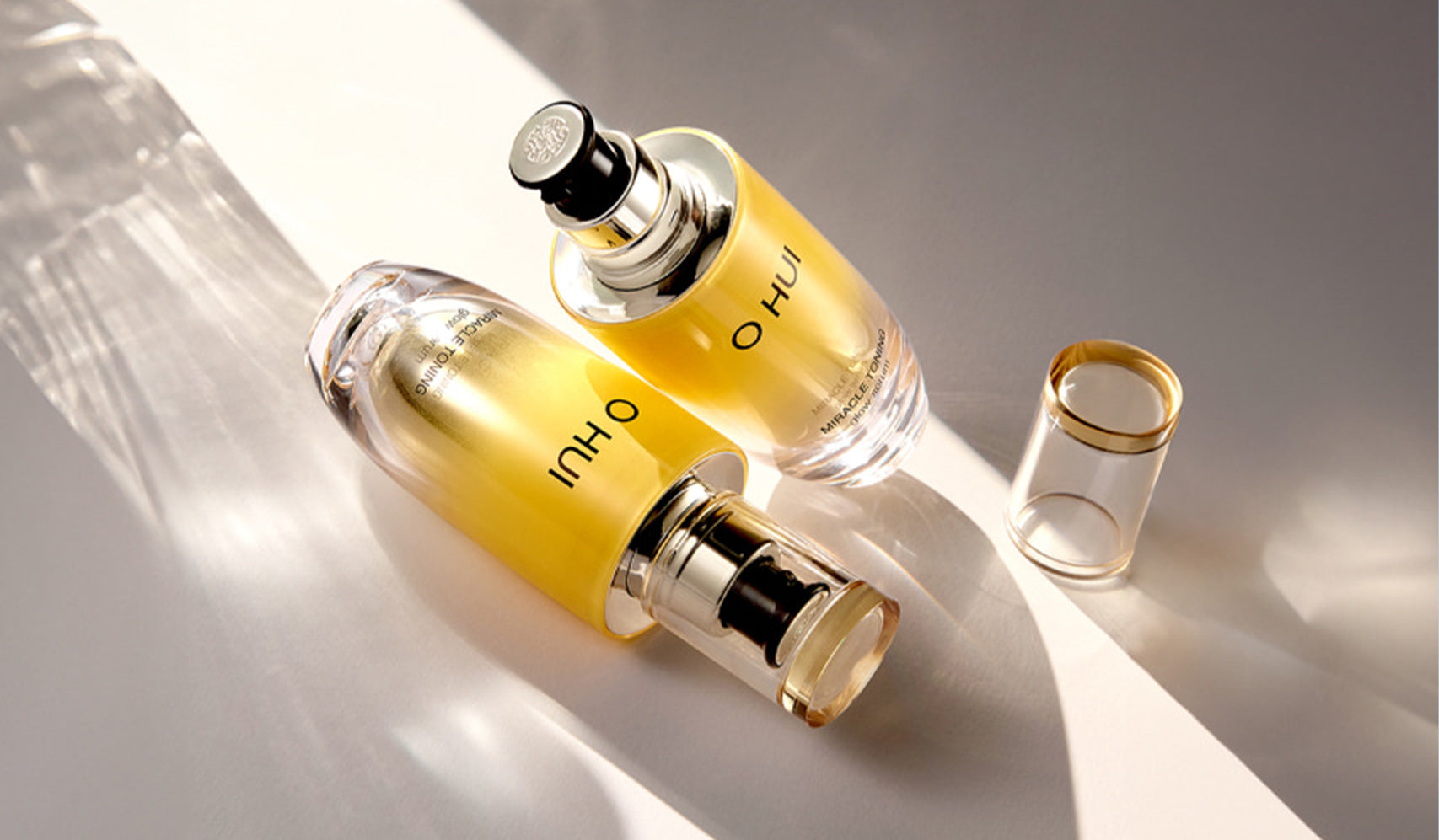 Two elegant bottles of O HUI skincare essence on a reflective surface with a golden cap and ambient lighting.