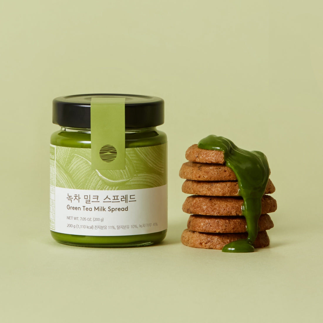Jar of OSULLOC Green Tea Milk Spread 200g next to stack of cookies topped with green tea milk spread on light green background.