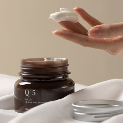 Hand applying QUA-T Dia All Green Total Essence Cream 100ml from open jar with lid on fabric.