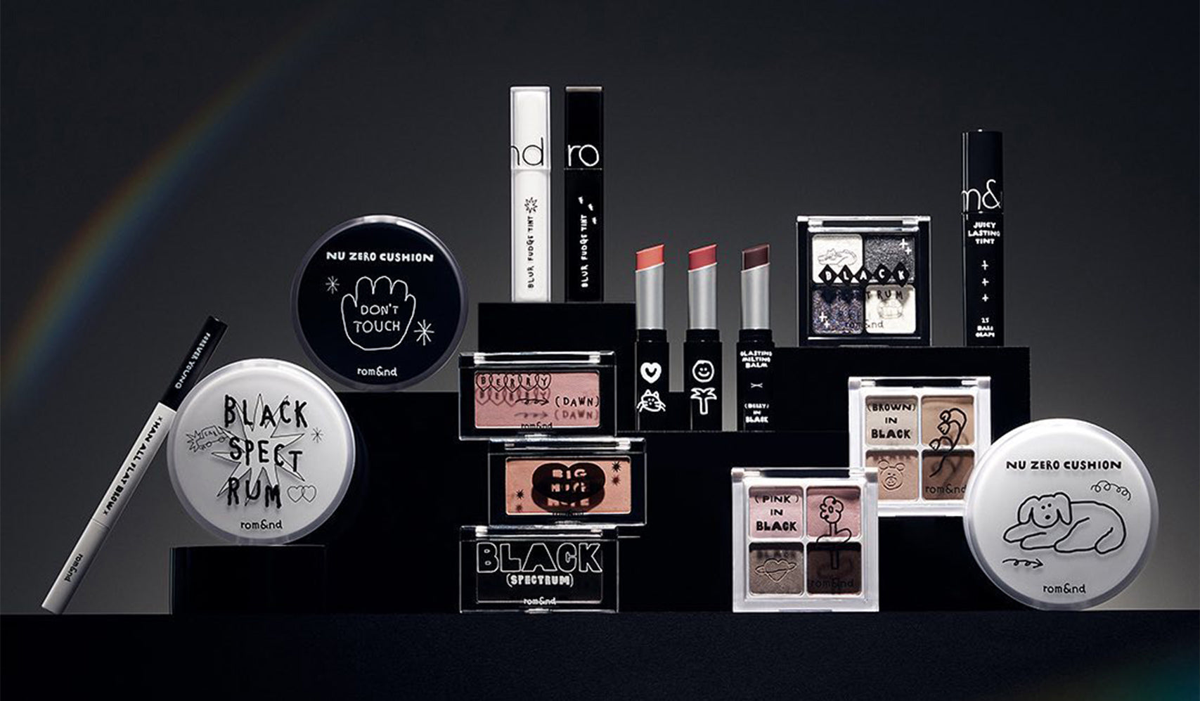 Collection of monochrome makeup products including lipsticks, eyeliners, and eyeshadows displayed on tiered black platforms.